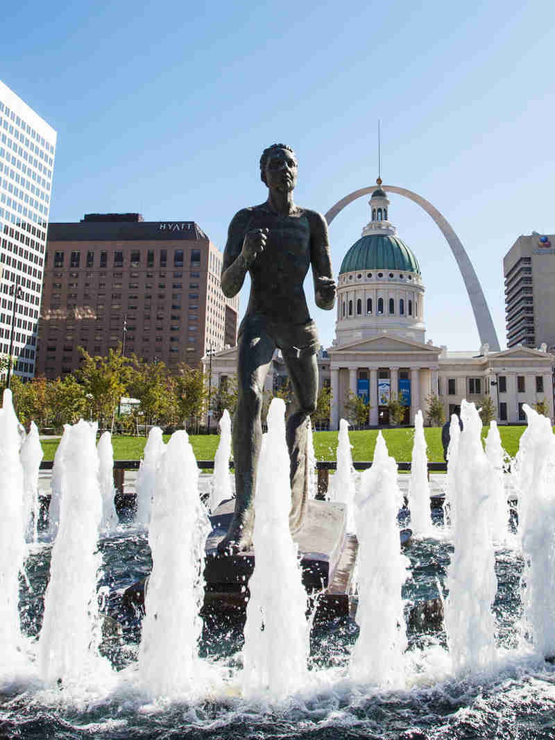 The Runner statue is splashed by the fountain in Kiener Plaza with the Old Courthouse and the Arch framing it in the background.
