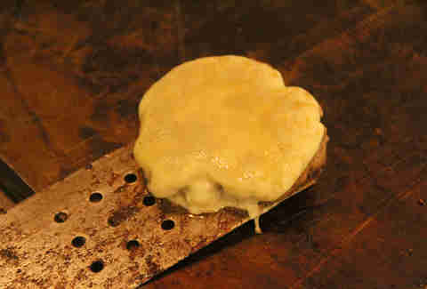 A spatula picks up PYT's provolone topped lasagna bun burger from the grill.