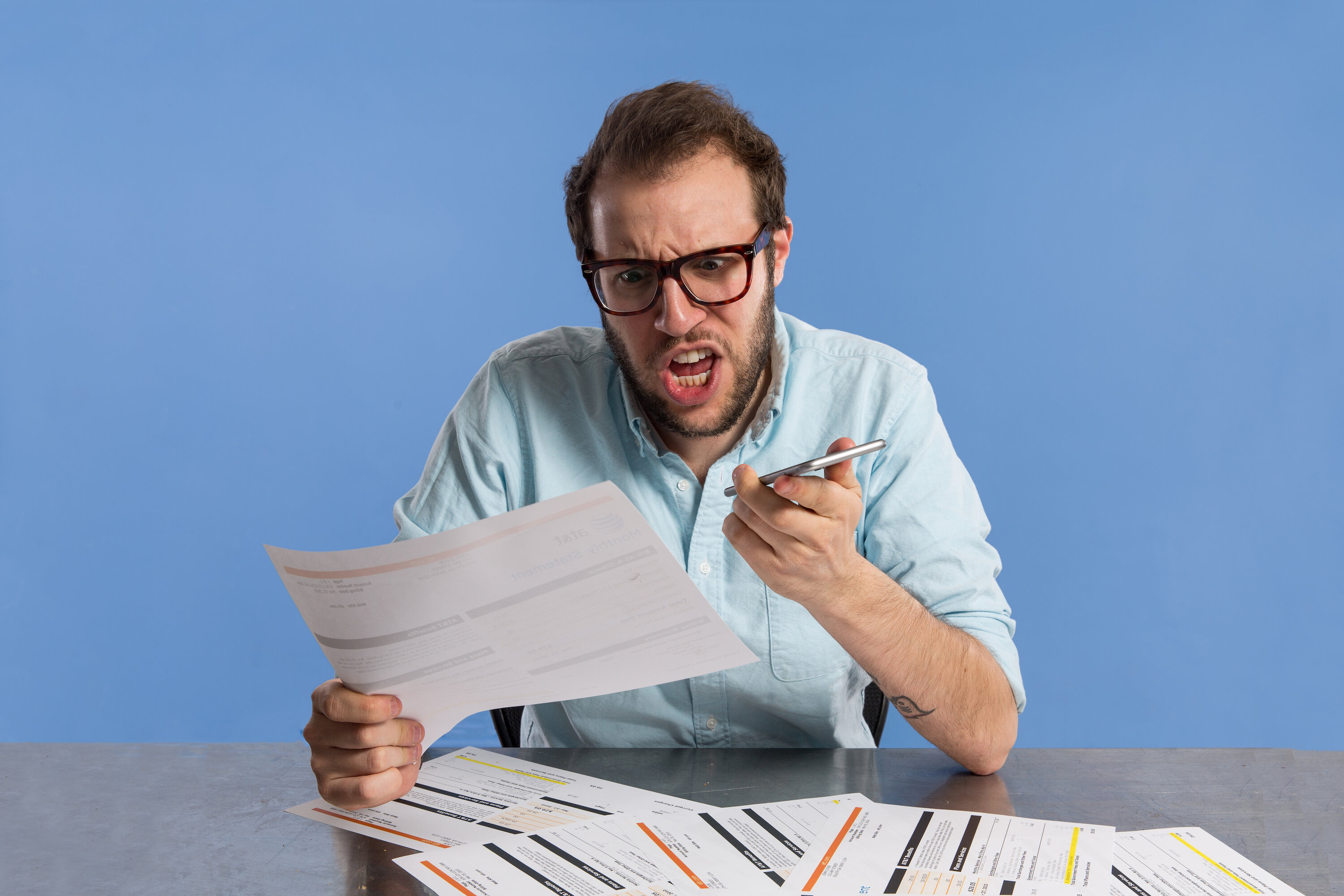 man yelling into iphone as he looks at a pile of bills