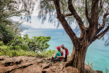 two women sitting beneath a tree on a cliff near the ocean