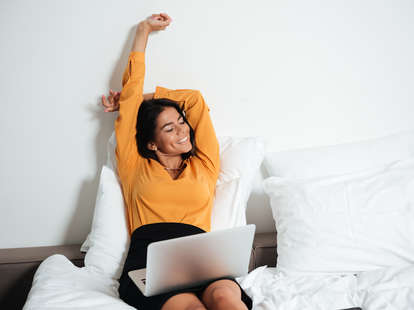 A woman sits up in bed, smiling with a laptop computer