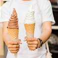 A person holds a chocolate soft serve ice cream cone and a vanilla soft serve ice cream cone.