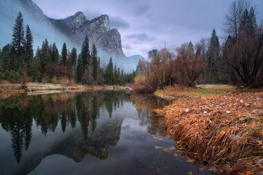 a river flowing toward trees and three mountain peaks