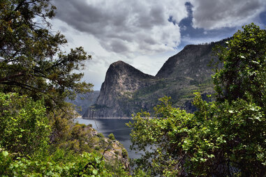 a lake near an enormous mountain cliff as seen through trees