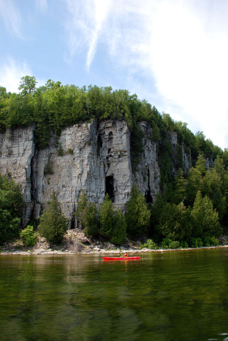 person canoeing down a river past a woodsy cliff