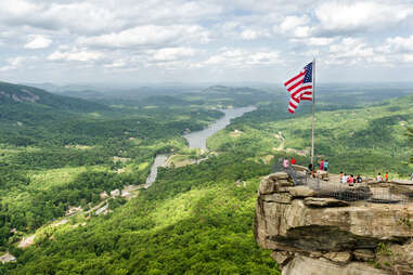 people on a cliff beneath an American flag overlooking sweeping forests and a river