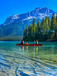 people canoeing on a crystal clear lake in front of mountains and a giant forest
