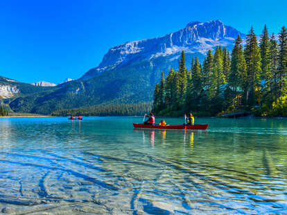 people canoeing on a crystal clear lake in front of mountains and a giant forest