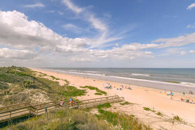 people walking down a small boardwalk path along a grassy sand dune to the beach