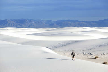 person walking across white sand dunes with mountains in the distance