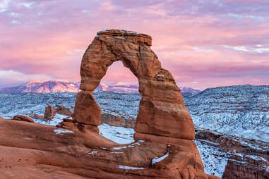 A colorful sunset lights up the La Sal Mountains behind iconic Delicate Arch in after a Febraury 2019 snowstorm in Arches National Park, Moab, Utah.