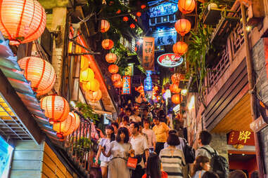 people going up crowded stairs on a busy, narrow street filled with lanterns and shops