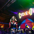 a drag queen wrestler on the edge of the ring with a neon sign reading chokehole in the background