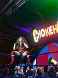 a drag queen wrestler on the edge of the ring with a neon sign reading chokehole in the background