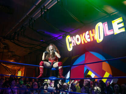 a drag queen wrestler on the edge of the ring with a neon sign reading chokehole in the background