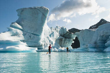 two adults on stand up paddle boards observe a hole melted in an iceberg