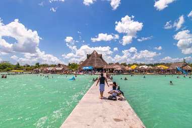 people on a long concrete boardwalk leading to a seaside village