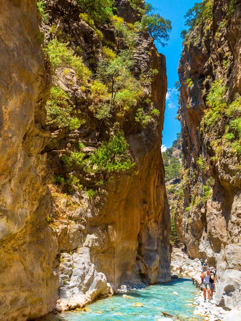People walk along a stream between giant rocks in the National Park Samaria Gorge