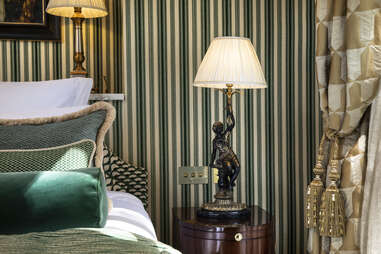 A bedside tabled with an elaborate lamp and rich green and gold wallpaper. 