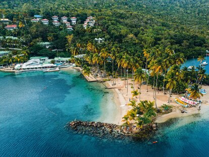 An aerial view of Marigot Bay in St. Lucia, which shows clear blue water, and a rocky shoreline covered in palm trees.