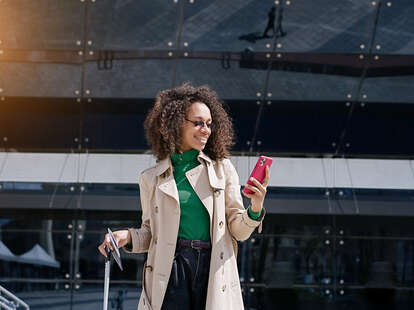 A woman waiting at the airport with phone in one hand and suitcase in the other.