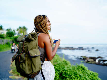 A young woman traveling with a backpack and a phone in her hand near a rocky shoreline.