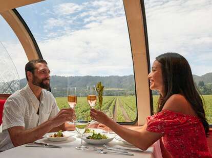 A couple sitting together in a glass domed train, sipping wine as they take the Napa Valley Wine Train through a vineyard. 