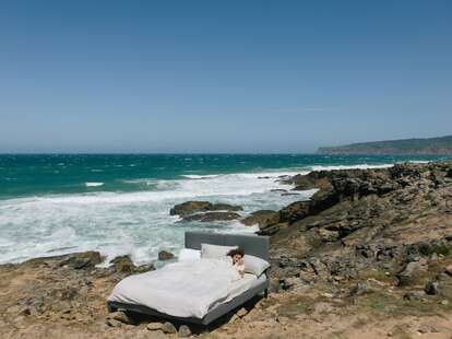 A woman sleeping in bed on the rocky shore of a beach.