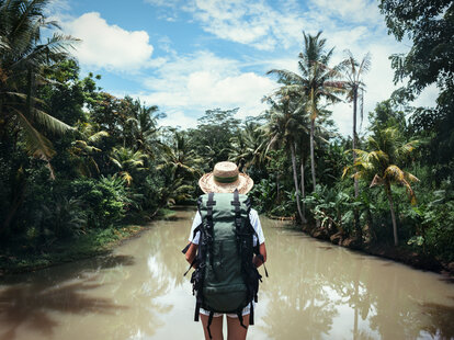 Traveling woman with backpack and straw hat looking at tropical river at sunny day.