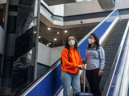 Women wearing masks while on escalator with suitcases.