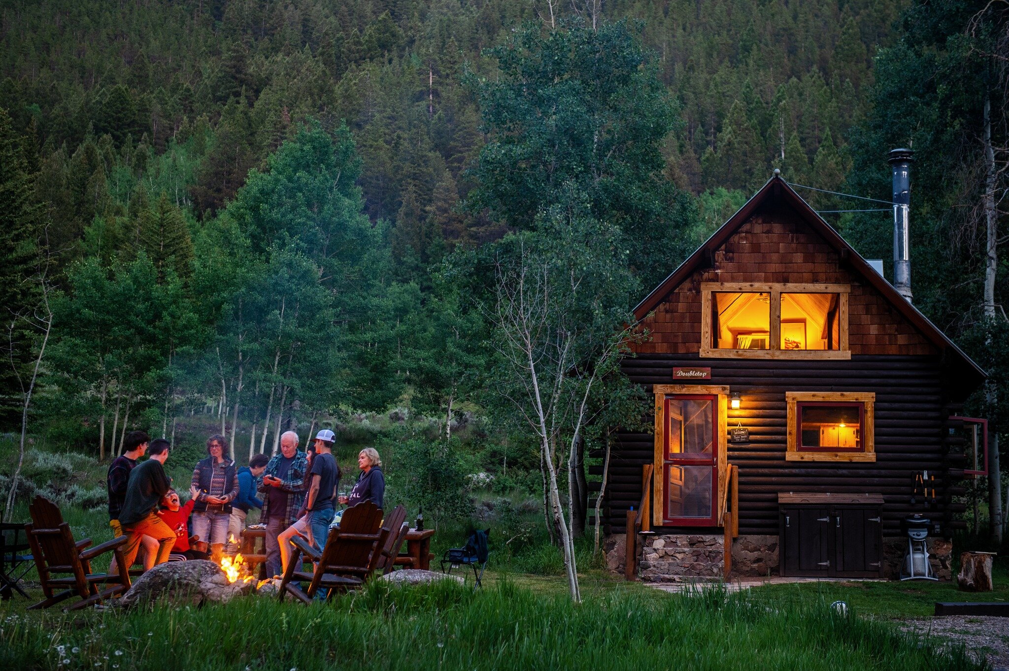 cabin in the woods at twilight next to group of people enjoying campfire