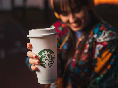 Modern young woman relaxing and listening to music with yellow headphones and drinking hot starbucks coffee in urban park during nice sunset.