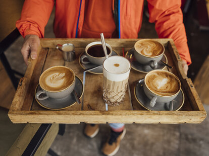 A man carries a wooden tray with different coffees on it in a cafe