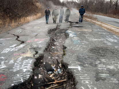 men walk down cracked highway with billowing smoke