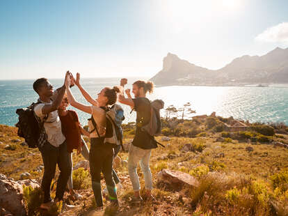 Young adult friends on a hike celebrate reaching a summit near the coast, full length, side view