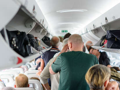 People are standing and sitting in an airplane cabin before disembarking.