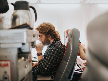 Man on a plane, reading a book, passing trolly with coffee.