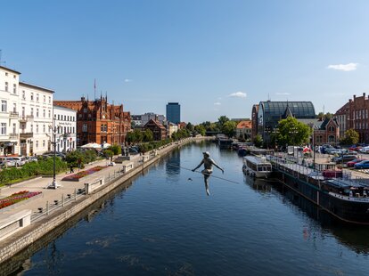 A view of the historic waterfront warehouses and buildings on the Brda River in downtown Bygdoszcz with the Crossing the River sculpture in the foreground.