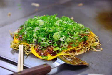 okonomiyaki, on a grill, covered in green onions, next to a spatula