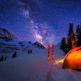 a glowing tent under the milky way with skis stuck in the snow outside of it