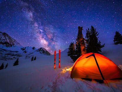 a glowing tent under the milky way with skis stuck in the snow outside of it