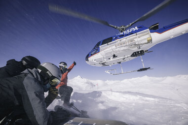 two heli-skiiers preparing to go down a mountain as the helicopter departs