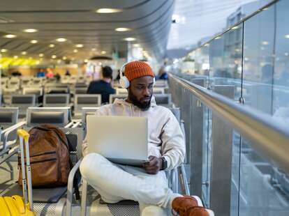 Man sitting in hall of airport terminal using laptop, wear headphones and listening music.