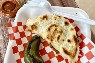a birria taco on red and white checked waxed paper alongside a pepper and a fork