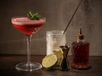 A martini glass filled with cherry juice, next to a decanter, a shot measurer, and a lime.