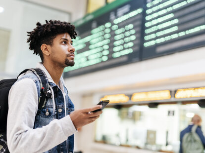 Young man at the airport looking at the list of destinations holding a cell phone.