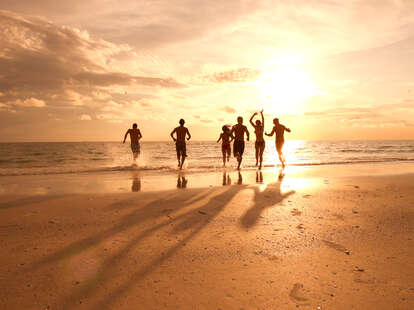Six young adults are running on the beach towards the water. The sky has a number of clouds, with the majority of the clouds on the left. The sun is low in the sky on the right half of the sky and is slightly covered with clouds.