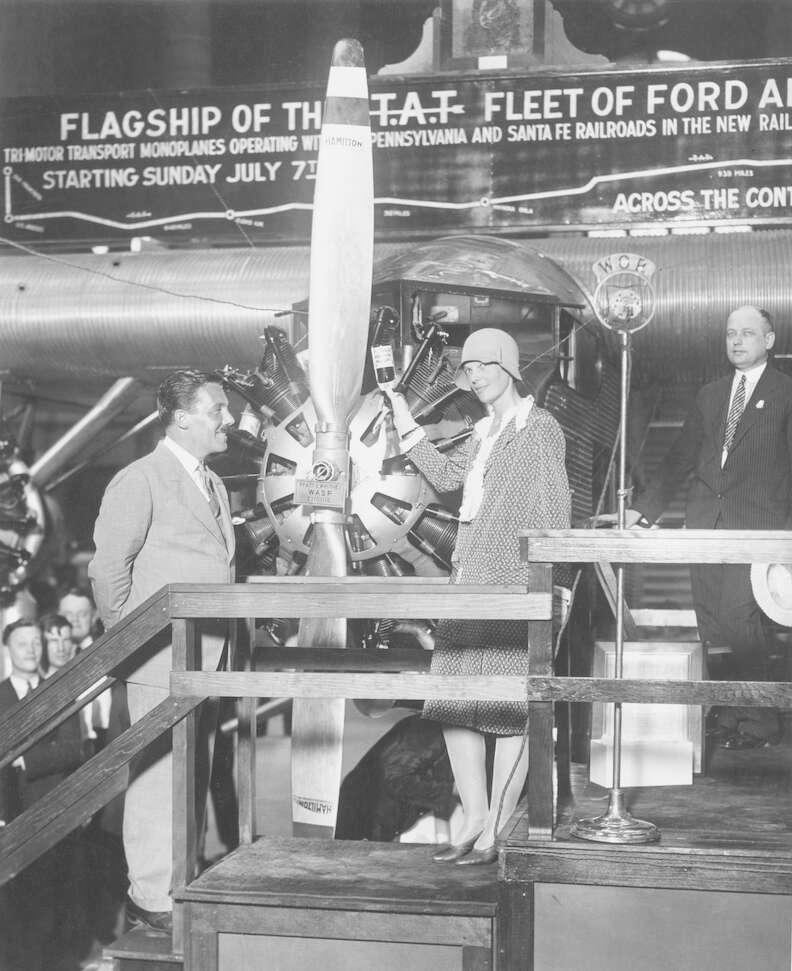 amelia earhart to the right of a tri-motor plane christening it with a glass bottle of ginger ale