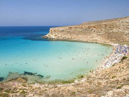 A View of Rabbit Beach or Spiaggia dei Conigli in Lampedusa, Italy. The beach is crescent shape, with crystal clear turquoise water.