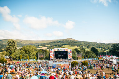 a crowd sitting on a wide lawn in front of a stage, with a mountain in the back